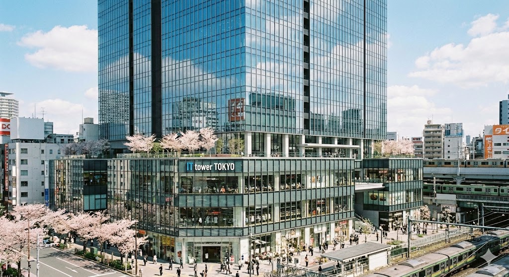池袋駅直結のIT tower TOKYOの外観、ガラス張りのモダンな建築、1階から4階の賑わう商業エリア、春の陽光、写実的